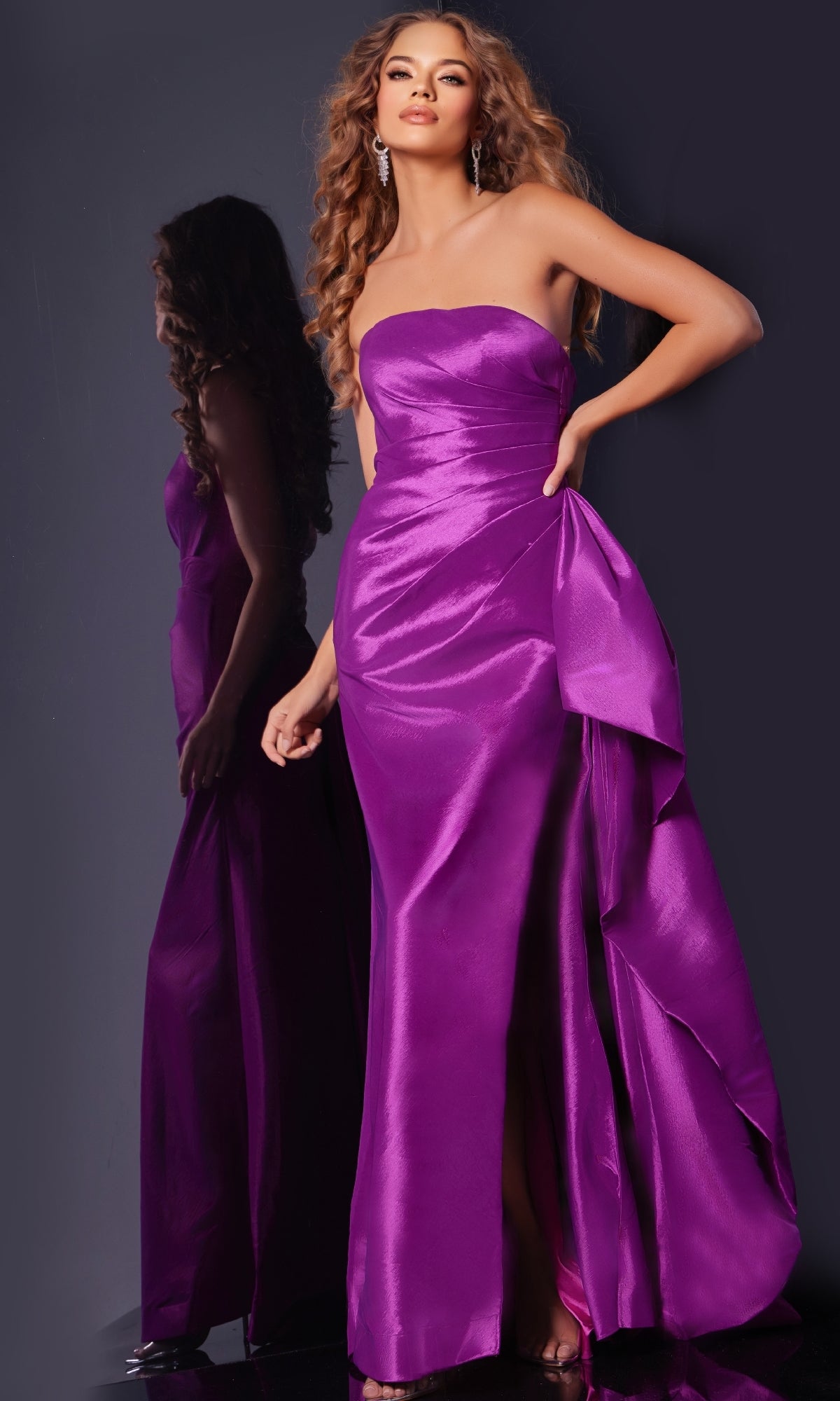 A woman poses with one hand on her hip, wearing the Long Prom Dress 36854 by Jovani—a fitted, strapless taffeta gown in shiny purple—standing before a dark background and a mirror reflecting her elegant look.